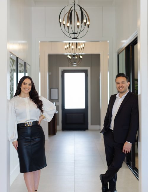 Two real estate advisors standing in a modern home entryway with chandelier, white walls, and dark front door.